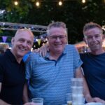 Three men pose together with arms around each other at an outdoor evening party, smiling under string lights and bunting with drinks on the table in front of them.