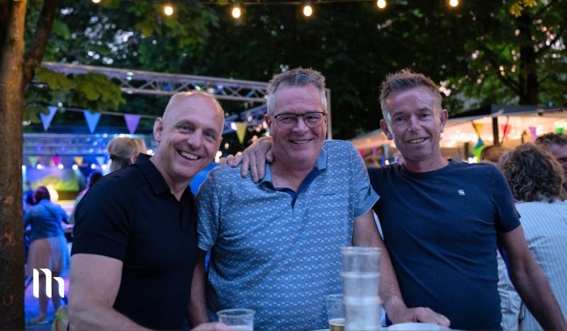 Three men pose together with arms around each other at an outdoor evening party, smiling under string lights and bunting with drinks on the table in front of them.