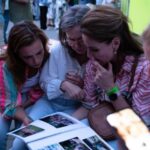 Four women sit closely together outdoors, looking at a photo album or magazine spread on a table. One woman uses a phone nearby.