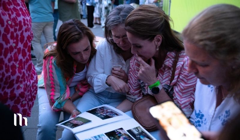 Four women sit closely together outdoors, looking at a photo album or magazine spread on a table. One woman uses a phone nearby.