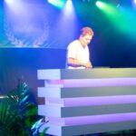 DJ performing on a stacked white booth under colorful stage lights at an outdoor event, greenery in the foreground
