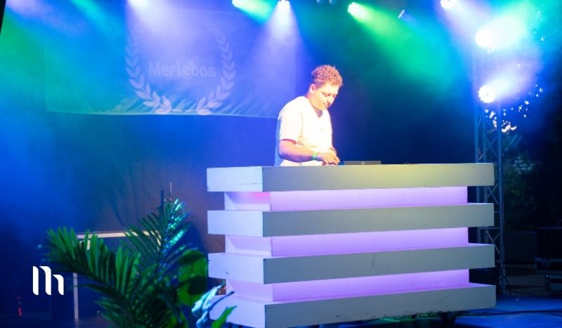 DJ performing on a stacked white booth under colorful stage lights at an outdoor event, greenery in the foreground