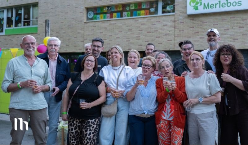 Group of about 15 adults posing together outside a brick building at a celebration, holding drinks with balloons and colorful banners in the background.