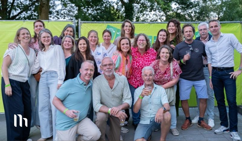 Group of diverse adults posing for a group photo at an outdoor event with a lime-green banner backdrop behind them showily smiling and holding drinks.