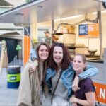 Three women smile and pose together in front of a bright orange food truck on a sunny day.