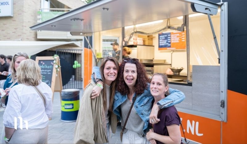 Three women smile and pose together in front of a bright orange food truck on a sunny day.