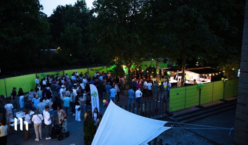 Evening outdoor festival with a crowd gathering around food stalls under trees and green barriers nearby.