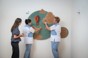Three people stand by a wall display featuring a colorful hot air balloon and fluffy clouds, two in light‑blue medical outfits and one in a patterned blouse observing.