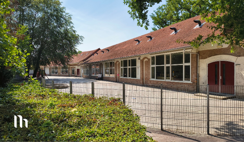 Row of brick school buildings with a red-tiled roof along a fenced courtyard on a sunny day.