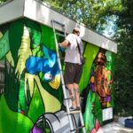 Graffiti artist on a step ladder painting a vibrant exterior wall mural outdoors.