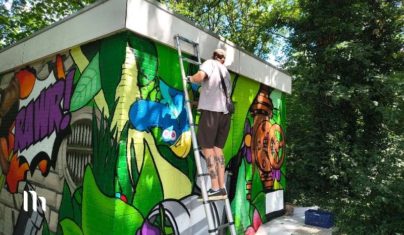 Graffiti artist on a step ladder painting a vibrant exterior wall mural outdoors.