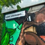 Colorful graffiti mural on a brick wall showing a brown dinosaur head with sharp teeth, green leaves on one side, and a white speech-bubble panel with text.