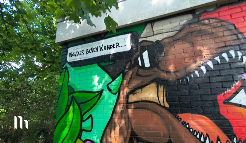 Colorful graffiti mural on a brick wall showing a brown dinosaur head with sharp teeth, green leaves on one side, and a white speech-bubble panel with text.