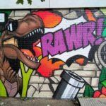 Colorful graffiti mural featuring a roaring T-rex with its mouth open, a bold purple 'RAWR!' burst, green plants, and a metal trash can on a brick wall behind it (urban street art). Includes the small text 'Wonder... Wonder' in a speech bubble at the top left.