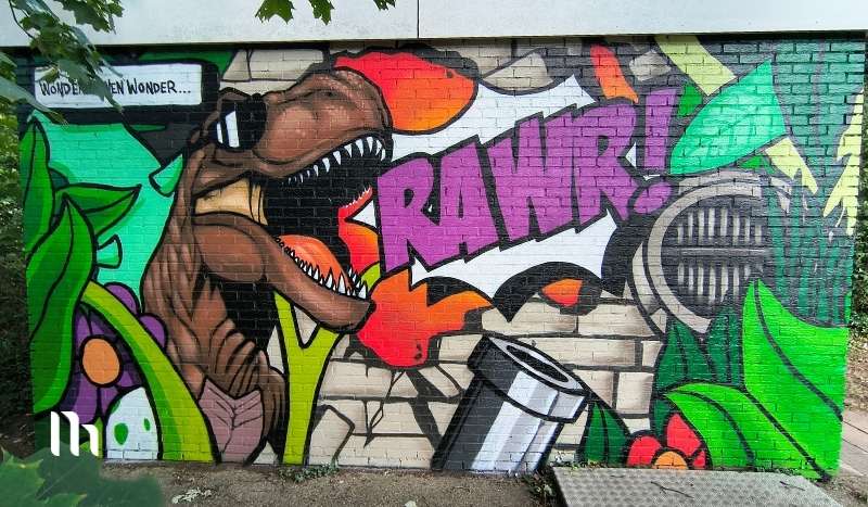 Colorful graffiti mural featuring a roaring T-rex with its mouth open, a bold purple 'RAWR!' burst, green plants, and a metal trash can on a brick wall behind it (urban street art). Includes the small text 'Wonder... Wonder' in a speech bubble at the top left.