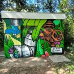 Colorful graffiti mural on a utility box in a leafy park, featuring pipes, a rusty valve, and green plants surrounding it.