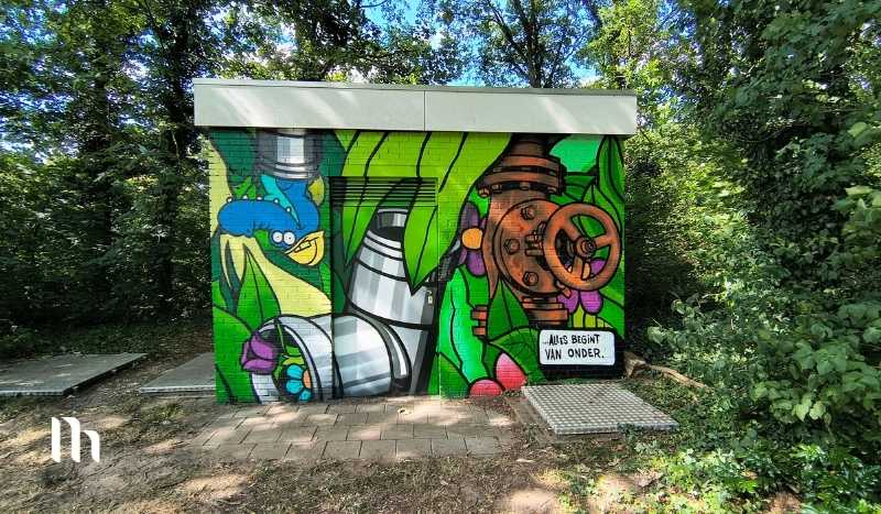 Colorful graffiti mural on a utility box in a leafy park, featuring pipes, a rusty valve, and green plants surrounding it.