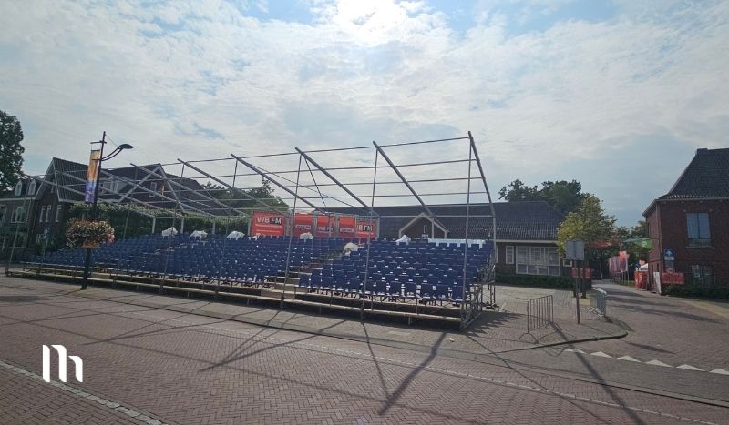 Outdoor temporary seating under a metal frame stage on a brick street, with banners in the background.